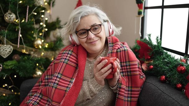 Mujer de mediana edad con cabello gris escuchando música bebiendo café sentado junto al árbol de navidad en casa - Foto, Imagen