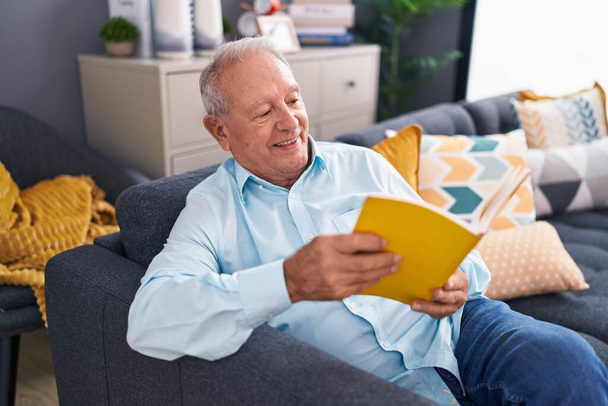 Середнього віку сіро-волохатий чоловік читає книгу, сидячи на дивані вдома
 - Фото, зображення