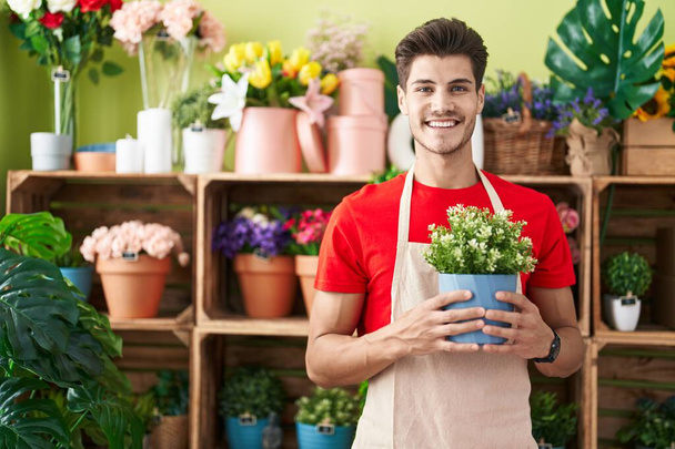 Młody Hiszpan pracujący w kwiaciarni, trzymający roślinę, wyglądającą pozytywnie i szczęśliwie, stojącą i uśmiechającą się z uśmiechem pokazującym zęby.  - Zdjęcie, obraz