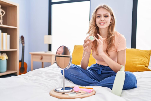 Молода блондинка застосовує лікування обличчя, сидячи на ліжку в спальні
 - Фото, зображення