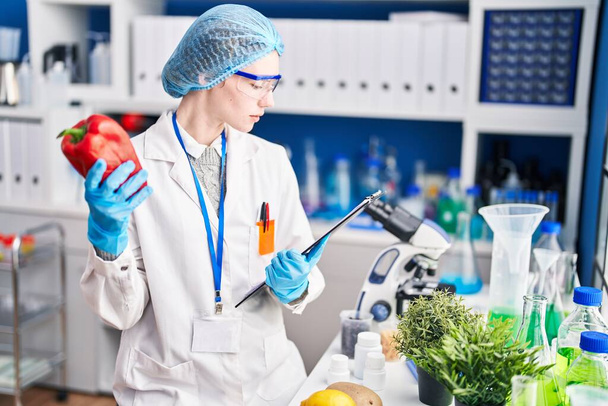 Jonge blonde vrouw wetenschapper met peper en checklist in het laboratorium - Foto, afbeelding