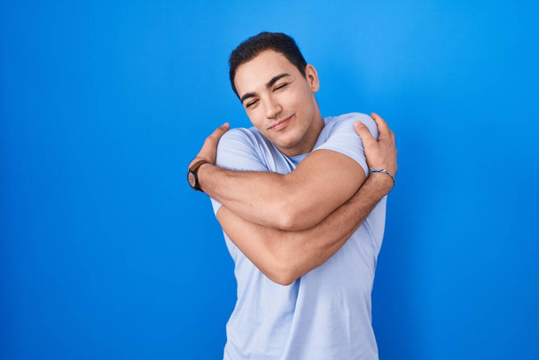 Junger hispanischer Mann, der vor blauem Hintergrund steht und sich glücklich und positiv umarmt, selbstbewusst lächelt. Selbstliebe und Selbstfürsorge  - Foto, Bild