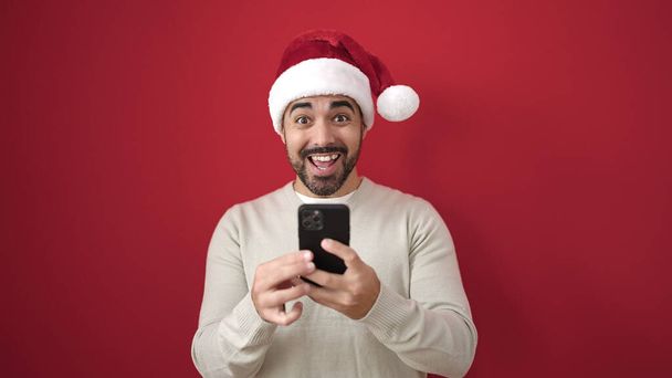 Mladý hispánec muž na sobě vánoční klobouk pomocí smartphone přes izolované červené pozadí - Fotografie, Obrázek