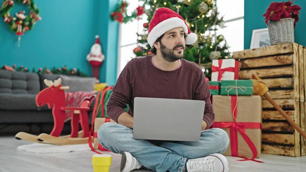 Young hispanic man using laptop drinking coffee celebrating christmas at home - Photo, Image