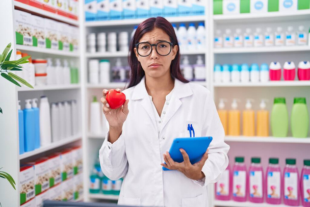 Young hispanic woman working at pharmacy drugstore holding red heart skeptic and nervous, frowning upset because of problem. negative person.  - Photo, Image