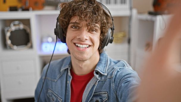 Usmívající se mladý muž s kudrnatými vlasy, sluchátky a džínovou bundou v moderním hudebním studiu. - Fotografie, Obrázek