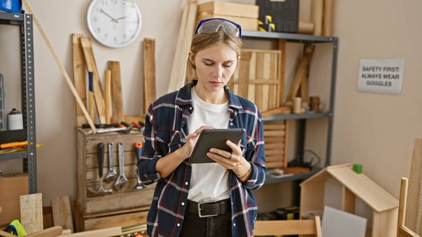 Odaklanmış genç bir kadın, iyi donanımlı bir atölyede marangoz aletleri ve güvenlik işaretleriyle tablet kullanır.. - Fotoğraf, Görsel