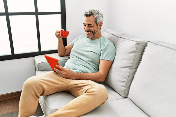 Middle age grey-haired man using touchpad drinking coffee sitting on sofa at home - Valokuva, kuva