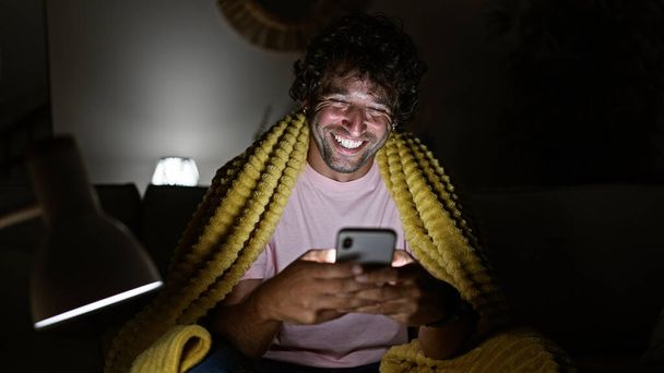 Veselý mladý hispánec si doma rád prohlíží smartphone, zabalený v útulné žluté přikrývce. - Fotografie, Obrázek