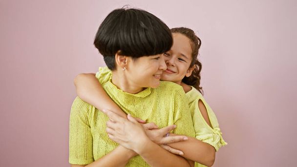 Zelfverzekerde moeder en dochter die een mooie, gelukkige knuffel delen terwijl ze glimlachen en samen staan over een geïsoleerde roze achtergrond, een casual levensstijl uitdrukken vol plezier, vreugde en positieve vibes. - Foto, afbeelding