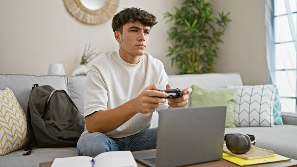 Junge hübsche hispanische Teenager, vertieft in ein intensives Online-Gaming-Match, zu Hause auf dem Wohnzimmersofa sitzend und mit dem Steuerknüppel jonglierend - Foto, Bild