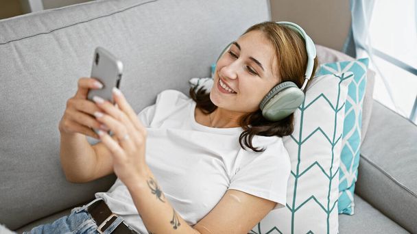 Νεαρή γυναίκα που χρησιμοποιεί smartphone φορώντας ακουστικά ξαπλωμένη στον καναπέ στο σπίτι - Φωτογραφία, εικόνα