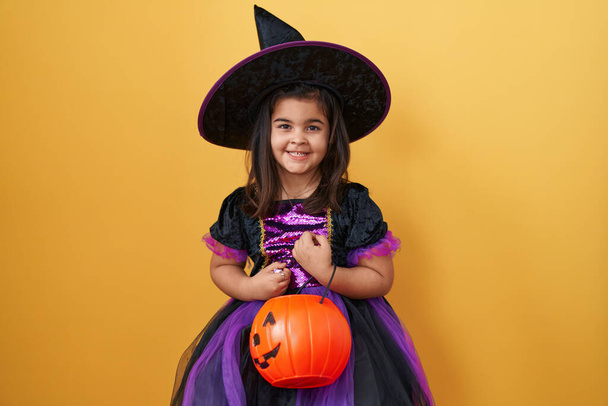 Entzückendes hispanisches Mädchen, das in ihrem Hexenkostüm positiv glücklich aussieht, selbstbewusst lächelt und sein zahmes Lächeln zur Schau stellt, hält einen Kürbiskorb vor isoliertem gelben Hintergrund. Halloween-Jubel! - Foto, Bild
