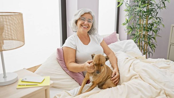 Eine lächelnde reife Frau mit grauen Haaren und Brille genießt die Zeit mit ihrem Hund auf einem bequemen Bett im Haus. - Foto, Bild