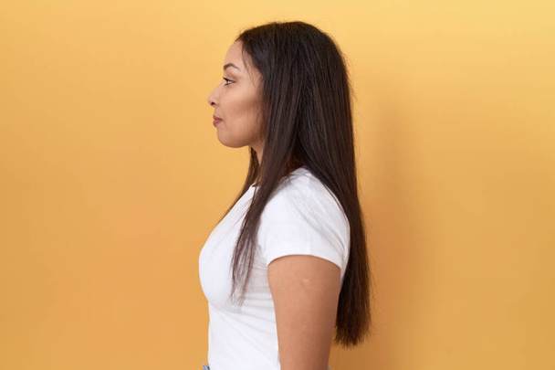 Jeune femme arabe portant un t-shirt blanc décontracté sur fond jaune regardant vers le côté, pose de profil relax avec le visage naturel et sourire confiant.  - Photo, image