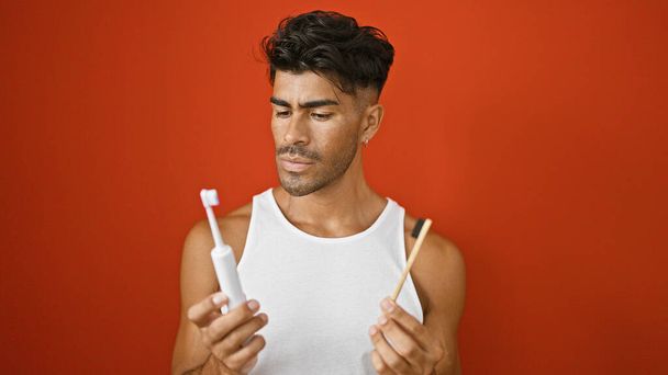 Jóképű spanyol férfi összekeveri a kézi és elektromos fogkefét a vörös fallal. - Fotó, kép