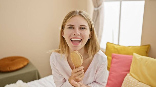 A joyful blonde woman with blue eyes holding a brush in a colorful bedroom setting, portraying a relaxed, cheerful moment at home. - Foto, Imagem