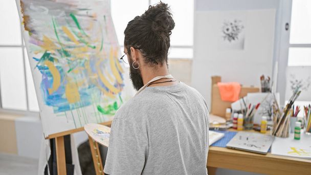 Όμορφος Ισπανός με γενειάδα σκέφτεται έναν πολύχρωμο πίνακα σε ένα στούντιο τέχνης.. - Φωτογραφία, εικόνα