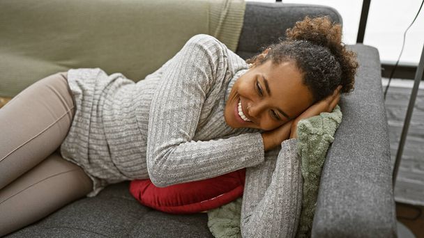 Расслабленная молодая женщина с вьющимися волосами, лежащая дома на диване, демонстрируя комфорт и счастье в уютном помещении - Фото, изображение