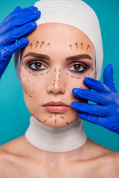 Muotokuva nainen plastiikkakirurgia potilas näyttää kosmetologi touch kasvot sininen lääketieteelliset käsineet yli syaani tausta. - Valokuva, kuva