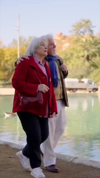 Pionowe wideo pary seniorów spacerujących razem na skraju sztucznego jeziora w miejskim parku - Materiał filmowy, wideo