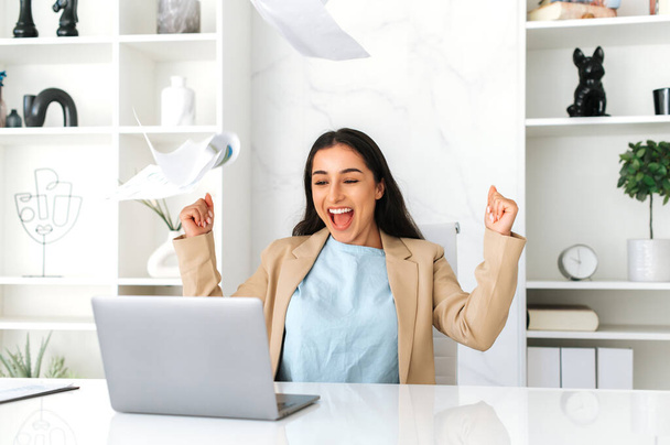 Joyful amazed happy indian or arabian business woman in elegant clothes, sit in modern office, rejoicing promotion at work, big profit, cheerfully throws documents, having fun, laughs, gesturing hands - Photo, Image