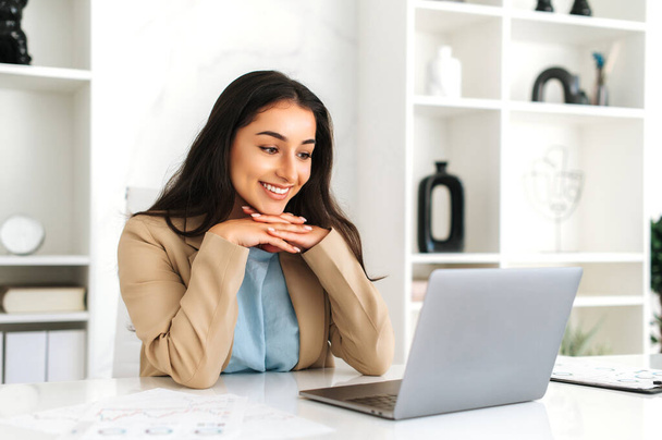 Pretty, attractive young adult indian or arabian business woman, multiracial entrepreneur, company employee, wearing suit, sitting in the modern office, looking at laptop screen and smiles - Photo, Image