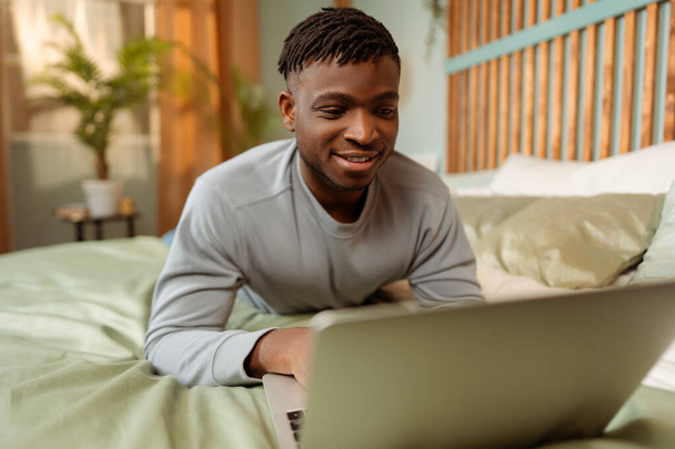 Portrét usmívajícího se atraktivního Afroameričana, ležícího na volné noze v posteli za použití notebooku, odlehlá práce doma. Technologický koncept - Fotografie, Obrázek