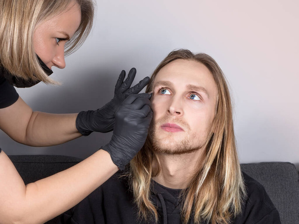 Real young man with blue eyes  during eyebrow correction. The eyebrow stylist shapes the client's eyebrows with tweezers.Cosmetology for men, professional skin and hair care - Photo, Image