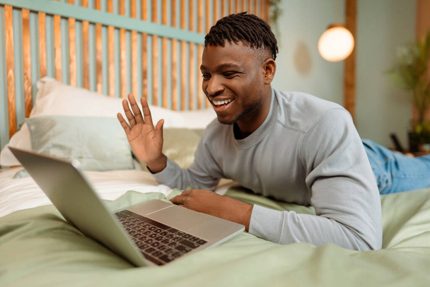 Porträt eines gutaussehenden, lächelnden afroamerikanischen Mannes, der zu Hause auf dem Bett liegt, seinen Laptop benutzt, Videoanrufe führt und mit der Hand winkt. Kommunikationskonzept - Foto, Bild