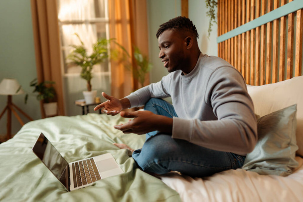 Retrato de un atractivo hombre afroamericano sonriente sentado en la cama en casa, usando una computadora portátil, haciendo gestos de videoconferencia, educación en línea. ? oncept de comunicación, tecnología - Foto, Imagen