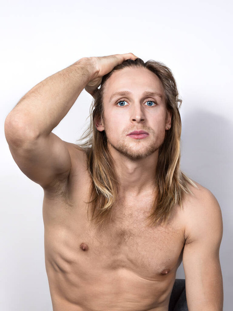 A handsome,seductive, topless young blond man with blue eyes sits on a chair,touching his long hair with his hand, and looks into the camera.Portrait of a real guy to the waist on a white  background - Photo, Image