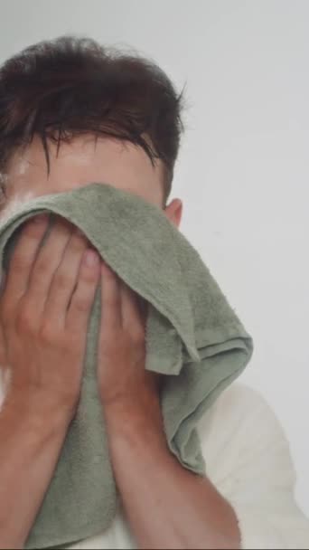 Vertical de hombre joven en albornoz blanco limpiando la cara con toalla delante del espejo en casa - Metraje, vídeo