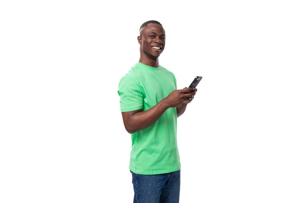 mladý šťastný přátelský Američan muž oblečený v zelené tričko a džíny chatování na telefonu. - Fotografie, Obrázek