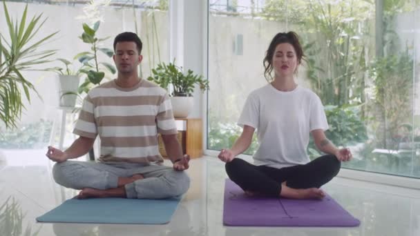 Pan shot de pareja biracial practicando yoga mientras se sienta en pose de loto en el suelo en salón con ventanas panorámicas - Imágenes, Vídeo