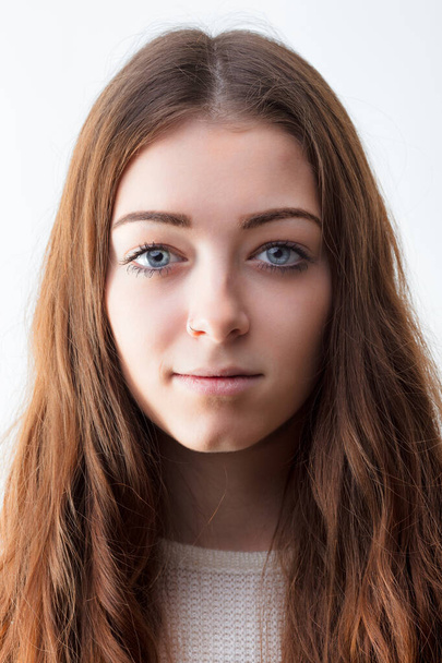 Jonge vrouw met een heldere huid en een neutrale uitdrukking staat tegen een witte achtergrond, haar lange bruine haar omlijst haar gezicht - Foto, afbeelding