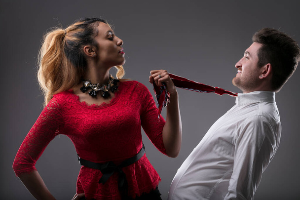 Woman in red with playful smirk holds a man's tie, suggesting control - Photo, Image