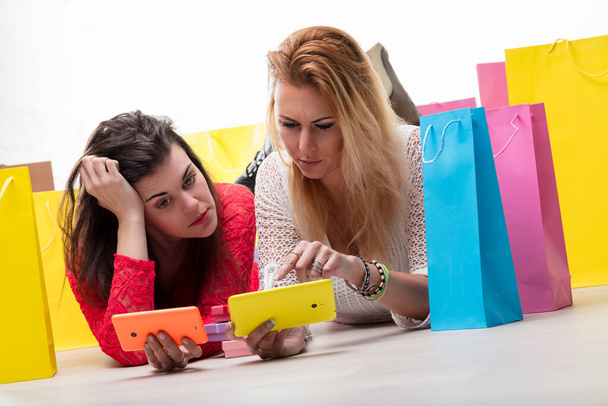 Two women enjoy a break after shopping, browsing on their phones amidst colorful bags - Photo, Image