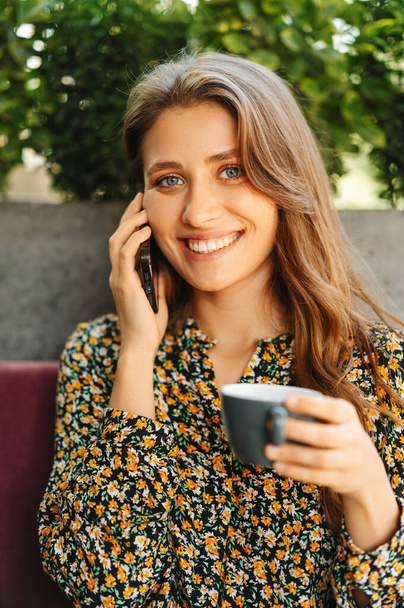 Vertical portrait of a positive young woman talking on the phone while sitting in a cafe. - Photo, Image