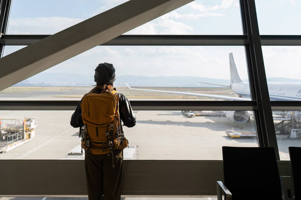 Matkustaja nainen suunnitelma ja reppu katso lentokone lento lentokentän lasi-ikkuna. - Valokuva, kuva