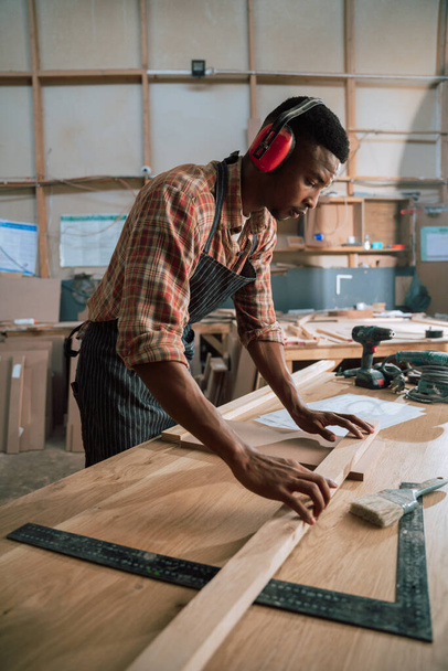 Jovem afro-americano na fábrica de carpintaria usa protetores auriculares enquanto trabalha. Foto de alta qualidade - Foto, Imagem