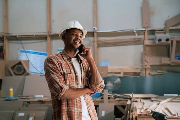 Jovem africano sorri enquanto fala ao telefone em um armazém de carpintaria. Foto de alta qualidade - Foto, Imagem