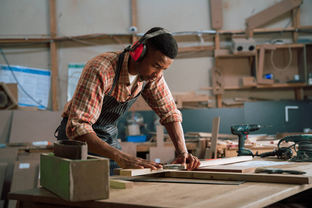 Skoncentrowany afrykański mężczyzna noszący nauszniki mierzy kawałek drewna na potrzeby projektu stolarskiego. Wysokiej jakości zdjęcie - Zdjęcie, obraz