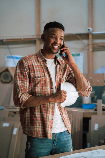 Šťastný Afroameričan v továrně na zpracování dřeva drží svůj klobouk a mluví po telefonu. Kvalitní fotografie - Fotografie, Obrázek