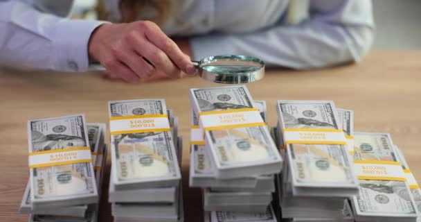 Banqueiro feminino examina cuidadosamente a designação em dinheiro através de lupa na mesa de madeira no escritório. Trabalhando como banqueiro e pilha de notas de dólar no banco - Filmagem, Vídeo