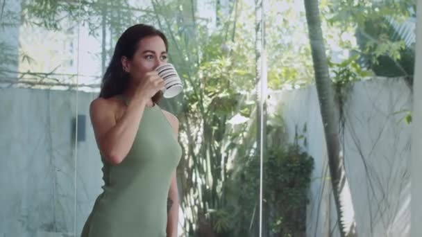 Inyección mediana de mujer atractiva joven en vestido de casa verde beber taza de té de hierbas mientras pasa el fin de semana en casa solo - Metraje, vídeo