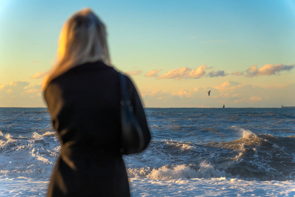 Блондинка в черном пальто смотрит на бурное море или океан на закате, избирательный фокус. Фото с задней стороны женщины, смотрящей на горизонт моря - Фото, изображение