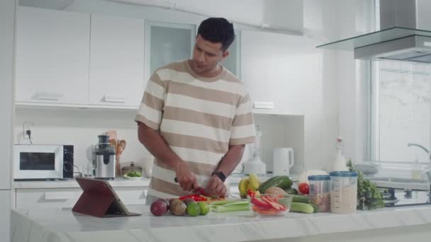 Medium shot of young Indian man looking at digital tablet while cooking salad for breakfast at kitchen - Footage, Video