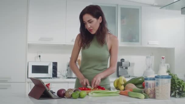 Tiro medio de mujer joven caucásica en ropa de casa verde haciendo ensalada de verduras mirando la receta en la tableta digital - Metraje, vídeo