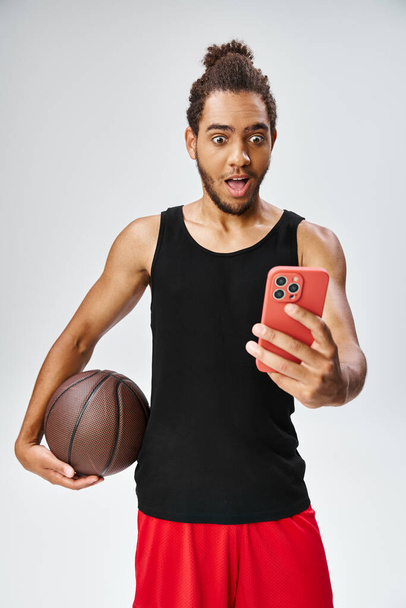 sorprendido hombre afroamericano en ropa deportiva con baloncesto mirando el teléfono, apuestas en línea - Foto, imagen
